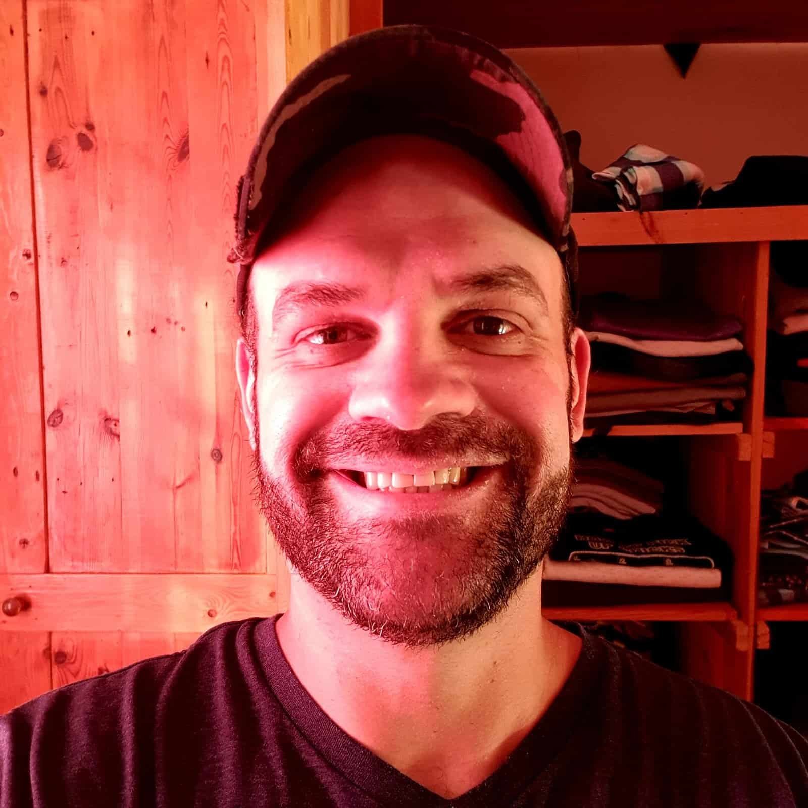 A joyful man wearing a cap, smiling warmly, in a cozy wooden room, representing faith and happiness in Christianity.
